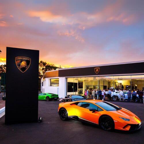 A yellow Lamborghini in front of a group of guests at a cocktail party mingling at sunset